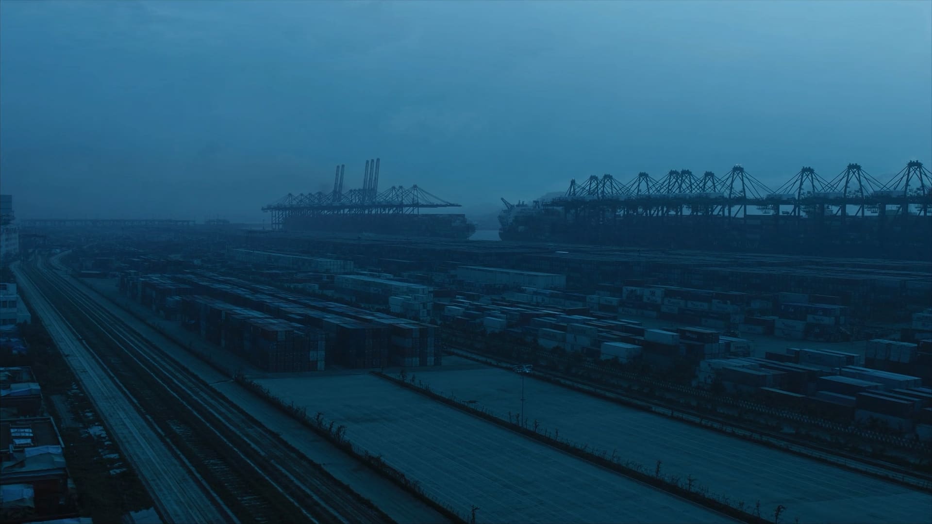 Shipping containers stacked in ordered rows across a container terminal at dusk, gantry cranes on the horizon